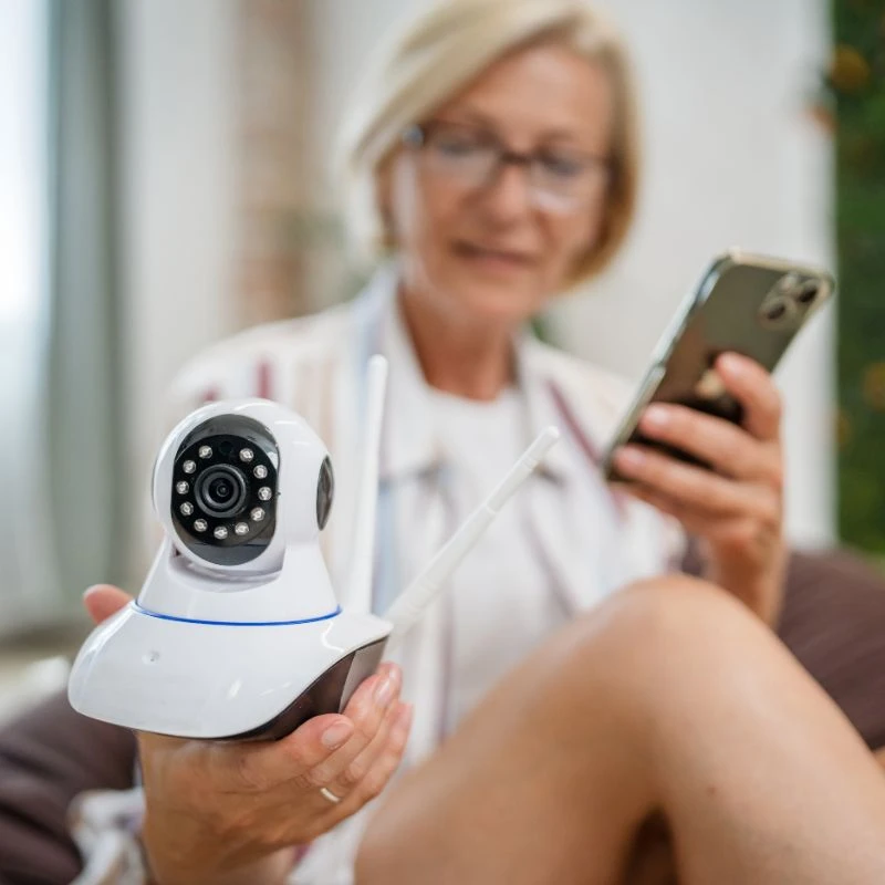 An elderly woman holding a smartphone while examining a home security camera, emphasizing home security systems.