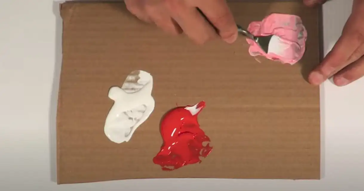 Mixing red and white paint on cardboard using a spoon, creating a hot pink color.