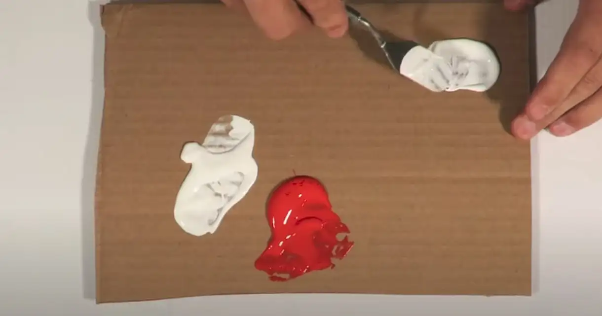 Artist preparing to mix white paint on a palette with a knife, ready to start with red paint nearby on cardboard surface.