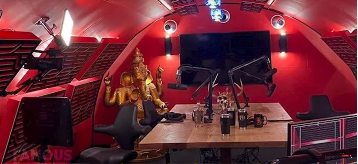 Interior view of the Joe Rogan Experience (JRE) red-themed podcast studio, showing a large wooden desk, professional microphones, ergonomic chairs, and a golden Ganesha statue.