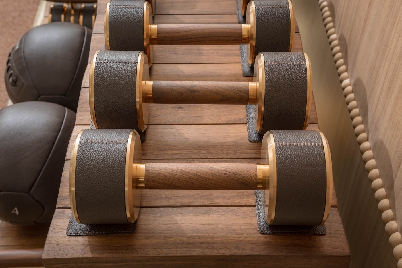 Luxury dumbbells with leather-wrapped ends and wooden handles on a polished wood surface, with leather medicine balls nearby