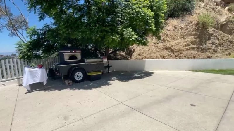 Outdoor patio of Jay Shetty house with concrete flooring, mobile pizza oven, and hillside backdrop