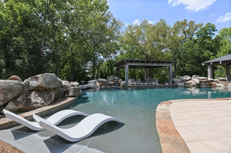 The lagoon-style pool at Bobby Bones' Nashville house with natural rock formations, a curved white lounge chair, a pergola with seating, and lush landscaping
