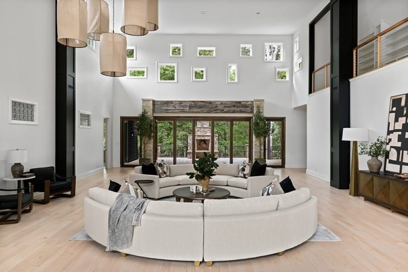 The living room in Bobby Bones' Nashville house with a curved white sectional sofa, stone fireplace, high ceilings with modern pendant lighting, and glass doors opening to the grounds