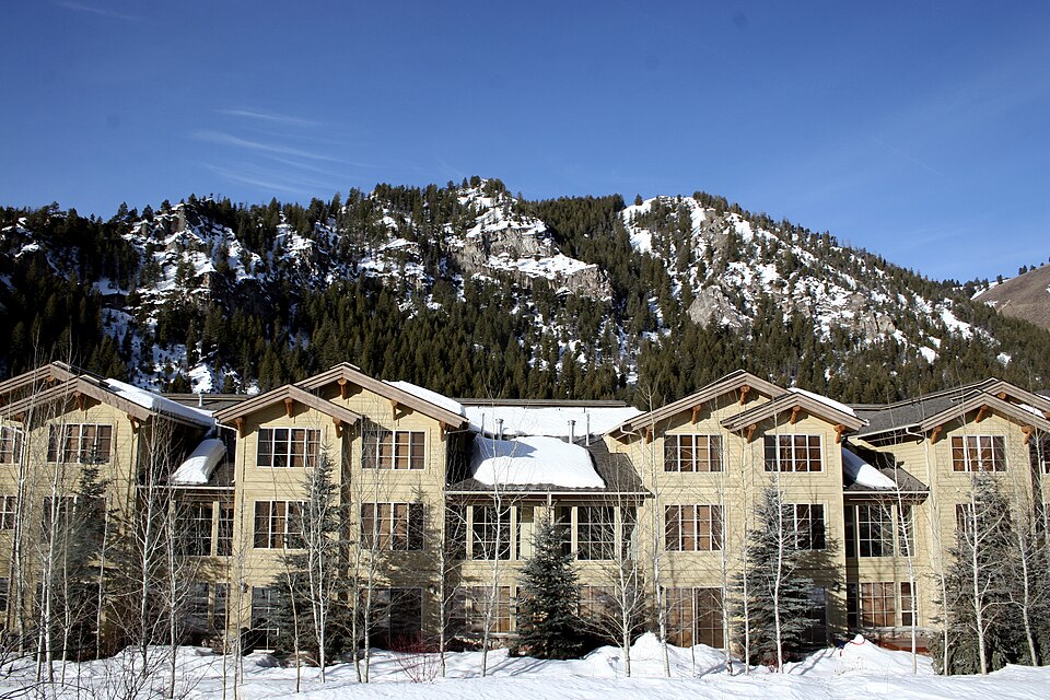 A beige multi-story building with snow-covered roofs stands before a forested, snow-dusted mountain under a clear blue sky.