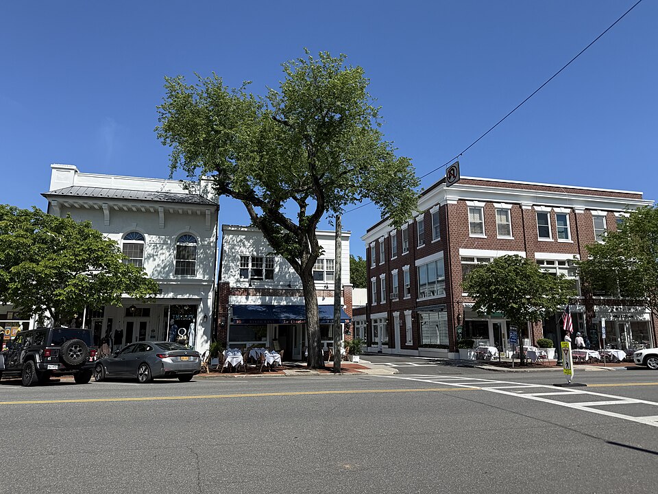 A sunny street corner with a white building, a brick building, parked cars, outdoor tables, and leafy trees under a clear blue sky.