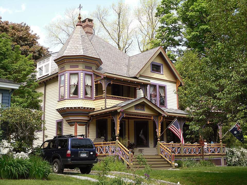 A Victorian-style house with a turret, wraparound porch, and American flag; a dark SUV parked on the lawn, surrounded by trees and greenery.