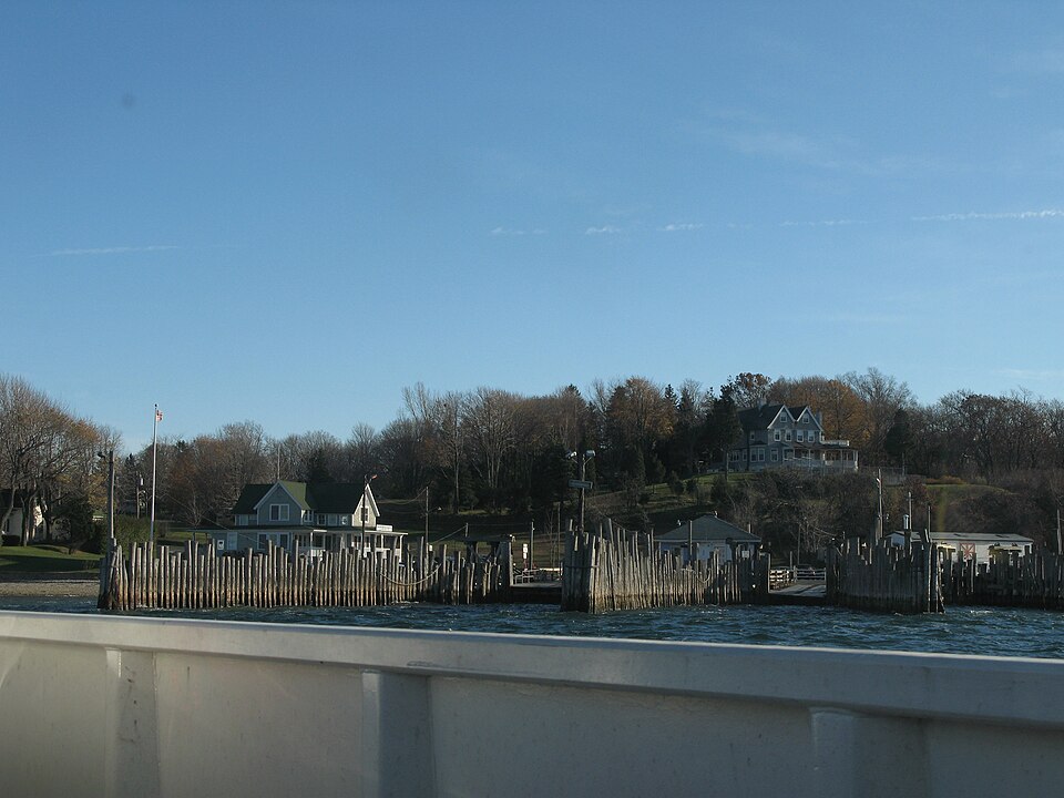 A waterfront scene with wooden pilings, houses on a hill, and a clear blue sky.